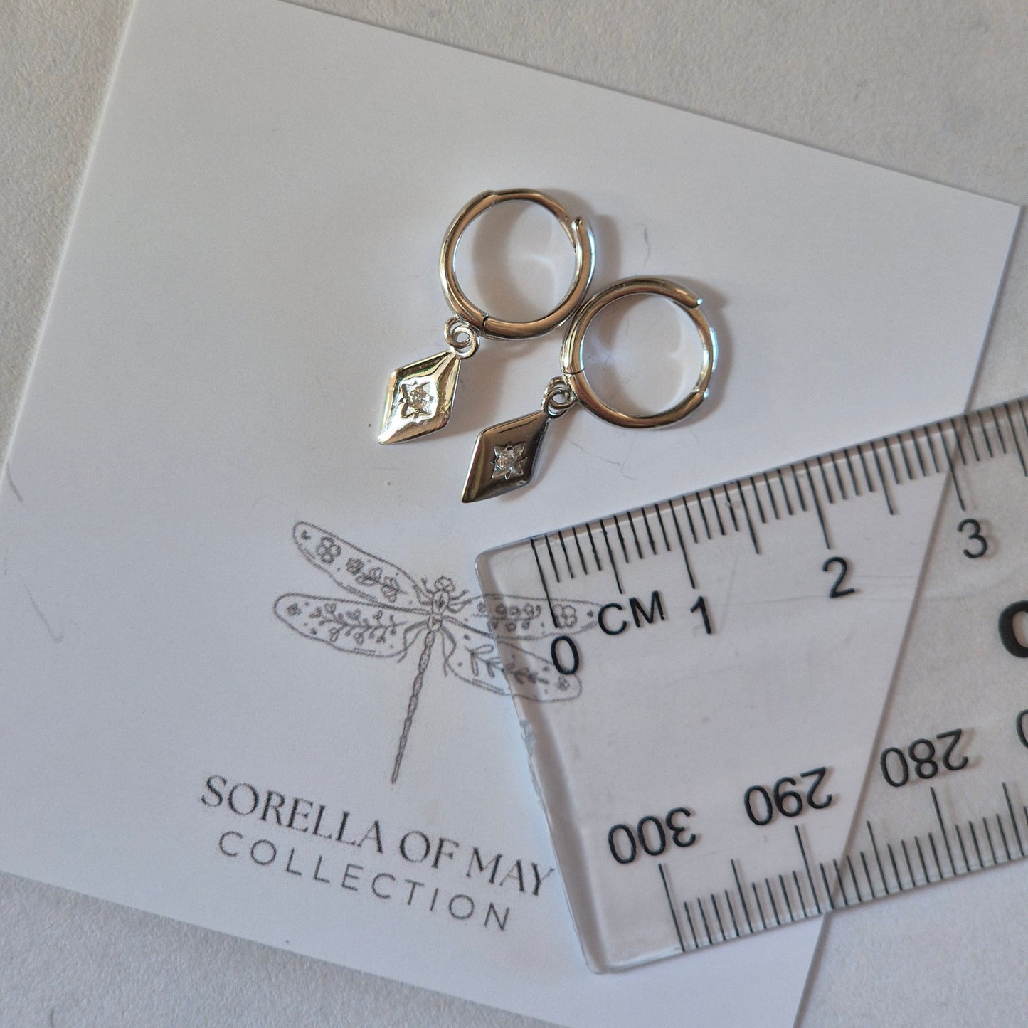 Silver earrings on a card with 'Sorella of May' branding, accompanied by a ruler for scale.