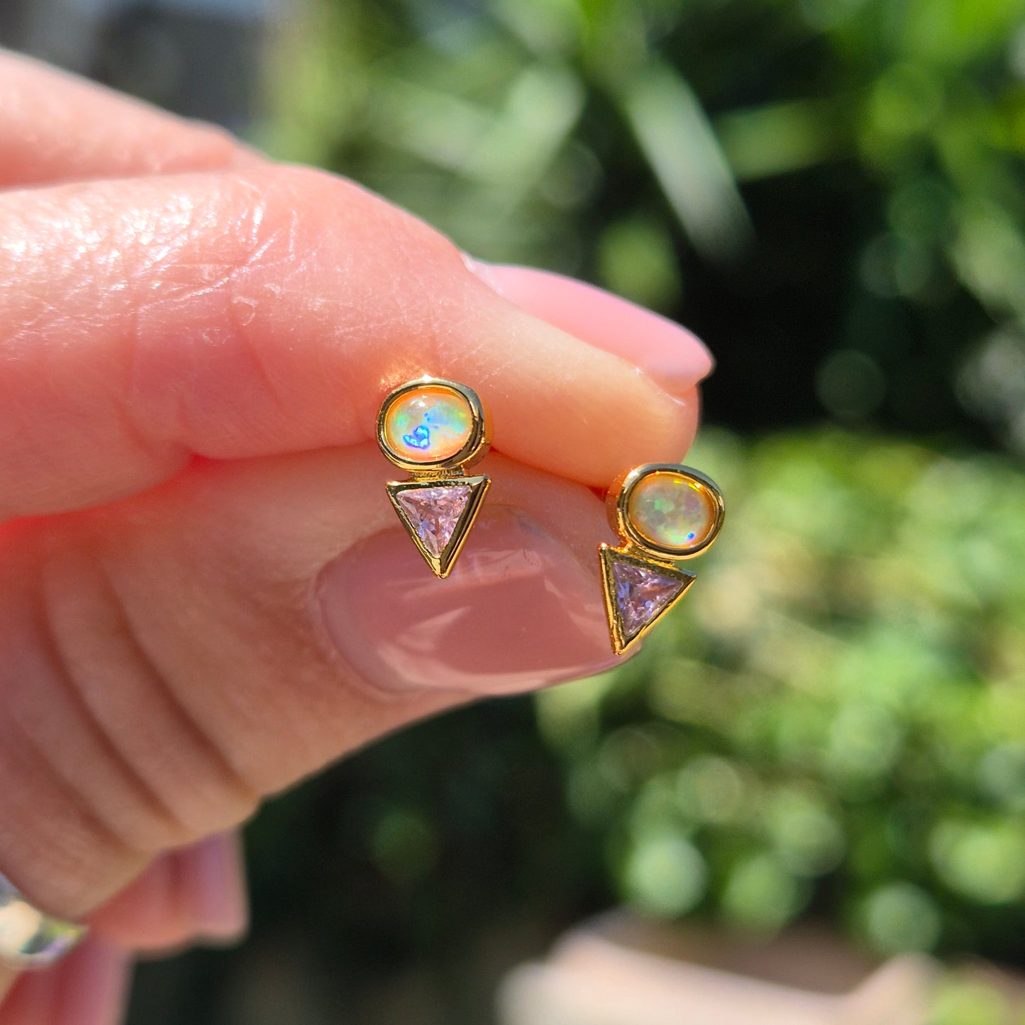 Gold Delta earrings with gemstones held between fingers against a blurred green background