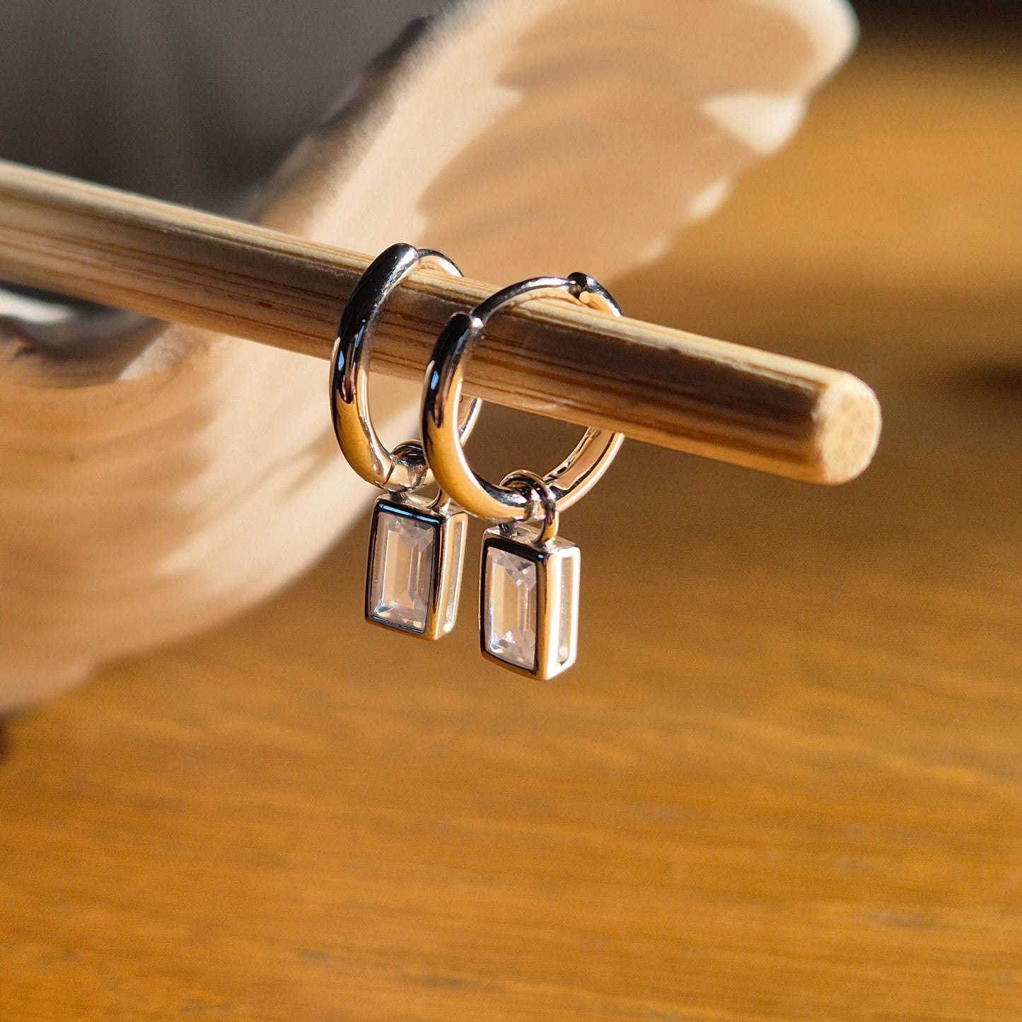 Close-up of Faye silver huggie earrings with Zirconia charms on a wooden stick on a blurred background