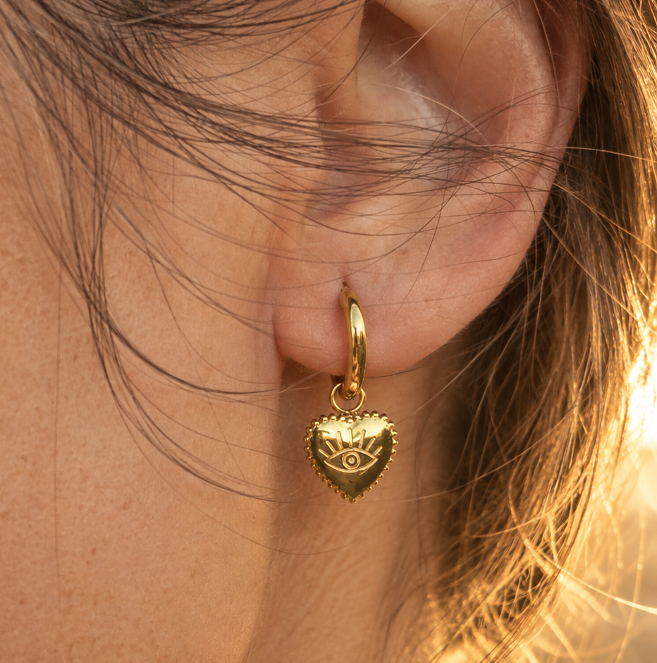 A close-up of a person's ear wearing a Sorella of May gold huggie hoop earring with a dangling heart charm featuring an evil eye design, lit by warm golden sunlight.