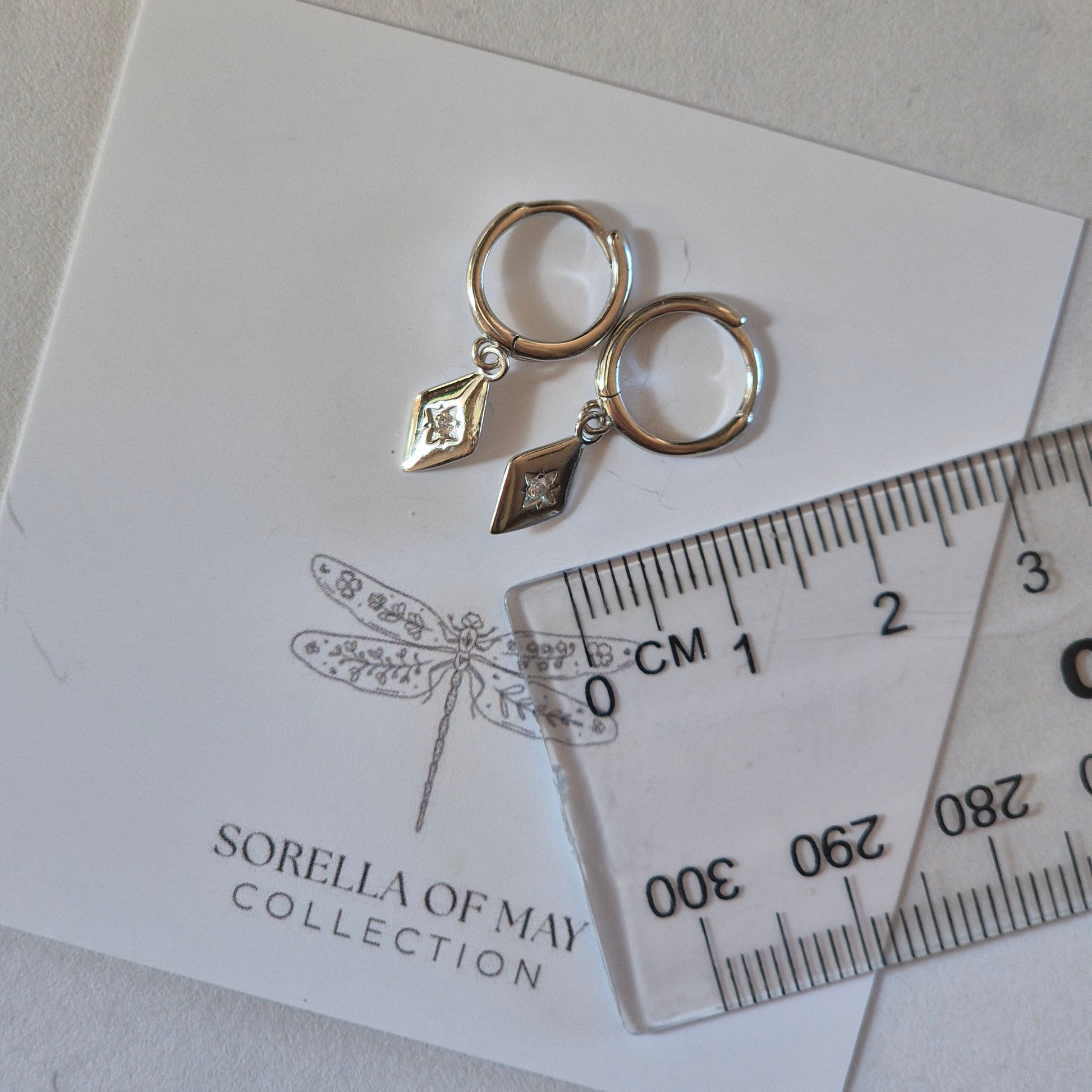 Silver earrings on a card with 'Sorella of May' branding, accompanied by a ruler for scale.