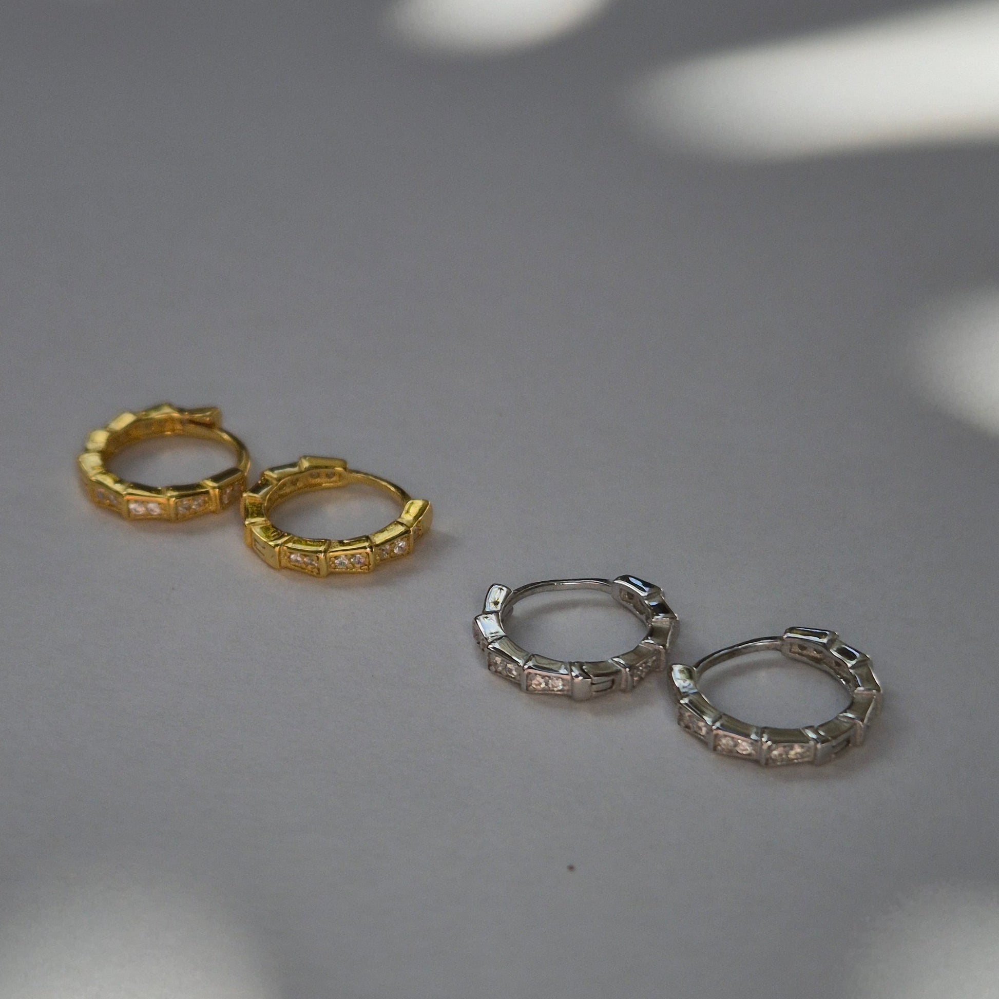 Two pairs of Frankie hoop earrings, one gold and one silver, on a gray surface with shadows.