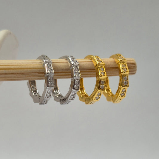 Two sets of Frankie hoop earrings, one silver and one gold, on a wooden stick against a gray background.