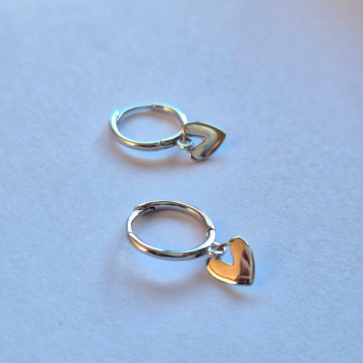 Two Sorella of May sterling silver hoop earrings, each with a dangling heart charm, one in polished silver and one in gold-tone metal, displayed on a light blue surface.