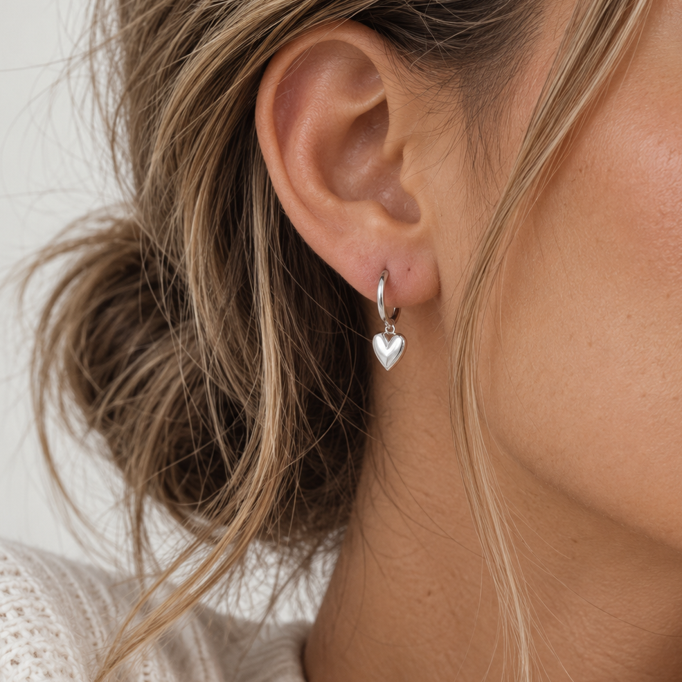 A close-up of a woman's ear wearing a Sorella of May sterling silver huggie hoop earring with a dangling polished silver heart charm.